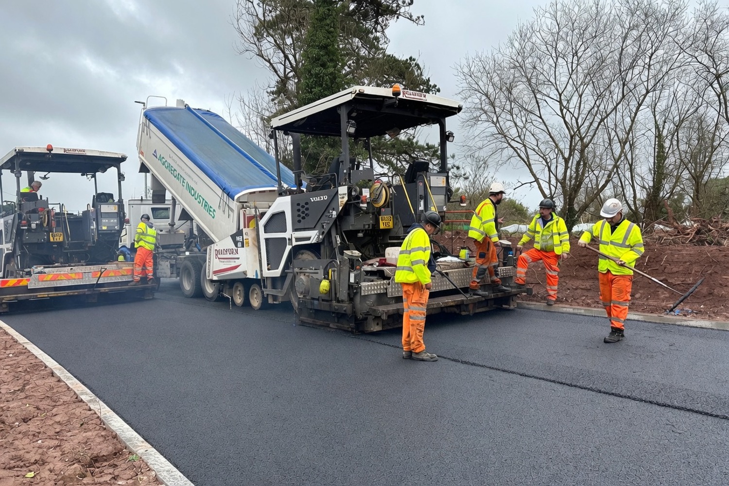 Paver fed by tipper truck, full-PPE crew laying a fresh asphalt mat