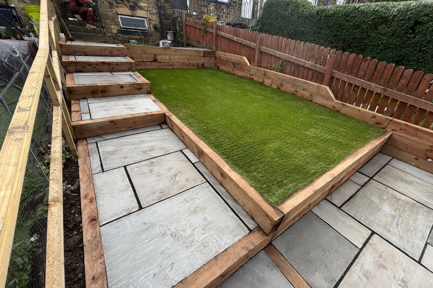 Sleeper-built tiered garden — sandstone-paved lower patio, sleeper-edged artificial-grass lawn, raised steps and pressure-treated timber fencing