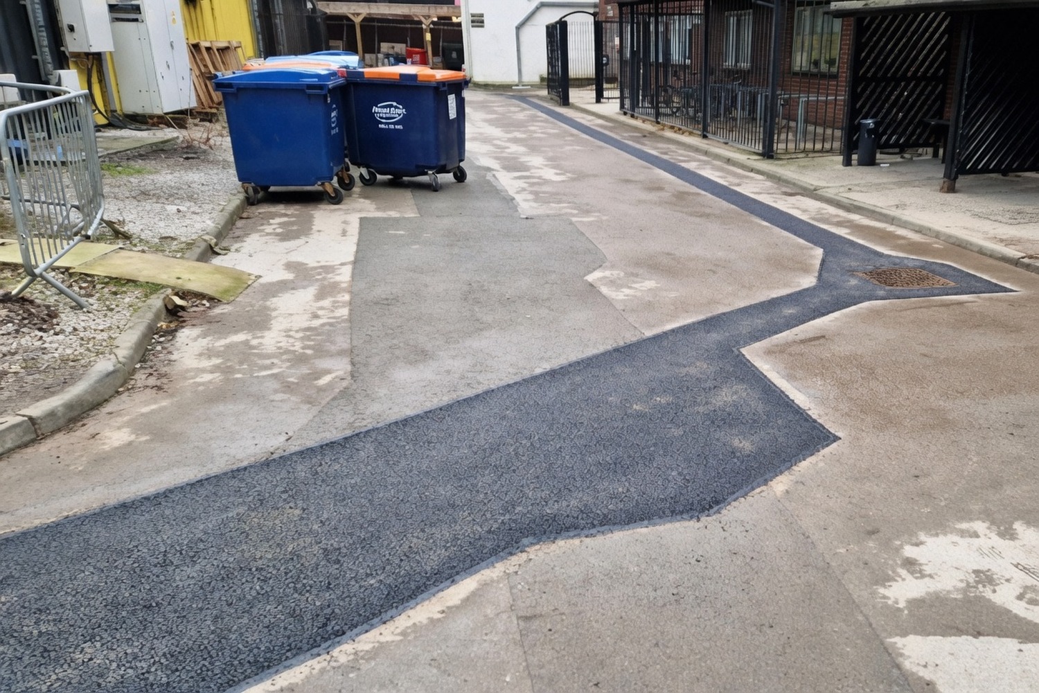 Long fresh asphalt reinstatement strip running along a kerb past wheelie bins
